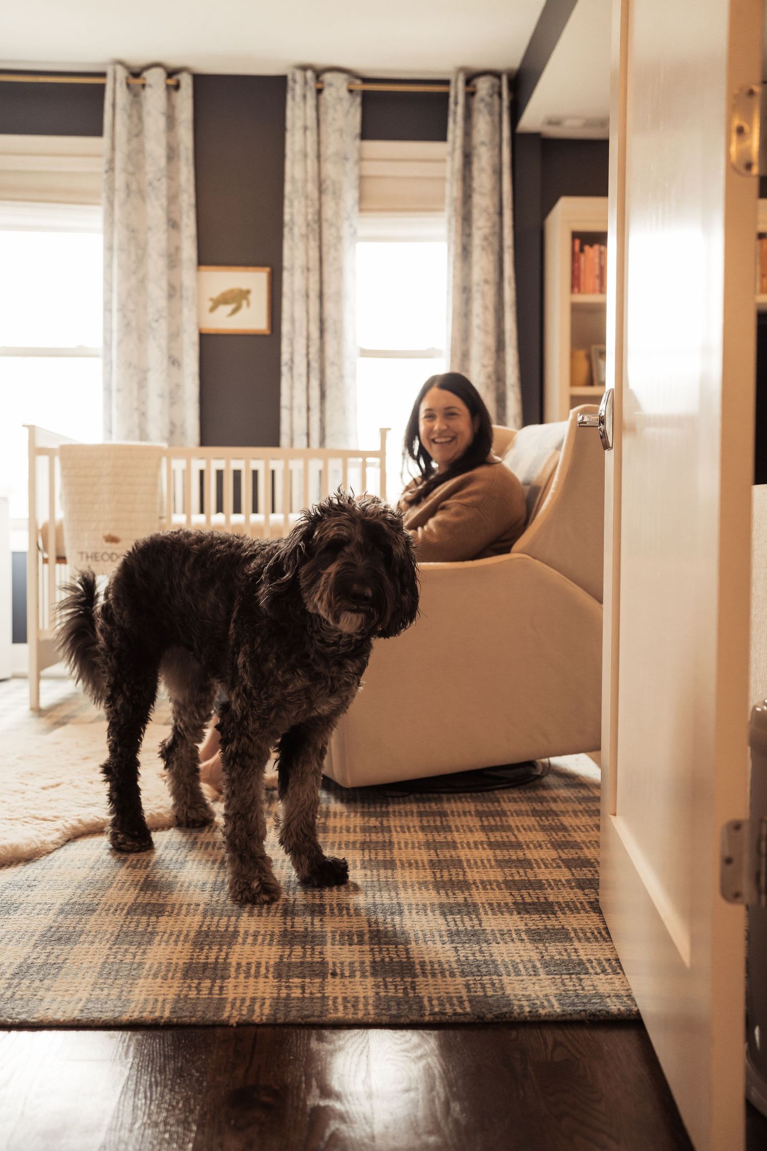 Woman sitting in a cream armchair smiles beside a large black fluffy dog in a navy blue nursery room.