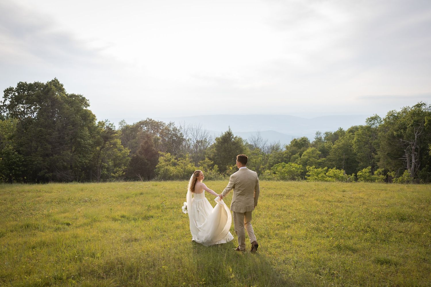 A bride and groom walk hand-in-hand through a lush green meadow surrounded by trees on their wedding day.