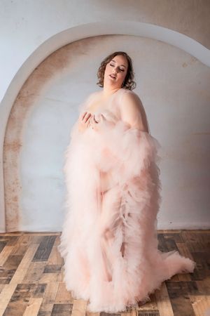 Elegant figure wearing a flowing blush pink tulle gown poses gracefully in front of an arched wall.