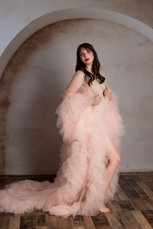 A figure in a flowing blush pink tulle gown poses elegantly against an arched wall and wooden floor.