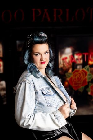 Rockabilly styled woman with blue hair wearing denim jacket poses against dark background with red neon signage.