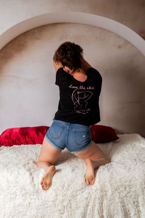 Someone wearing a black graphic t-shirt and denim shorts sits on a white furry surface with red pillows in an alcove.