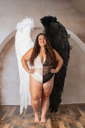 A boudoir photoshoot featuring contrasting black and white angel wings against a rustic wooden floor and neutral backdrop.