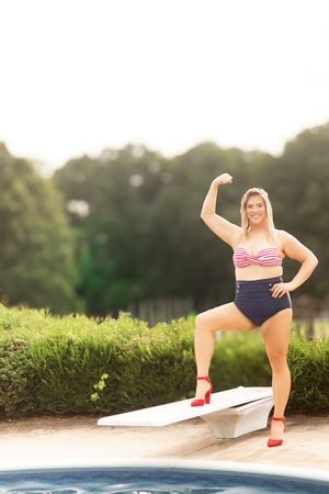 Woman in striped bikini top and high-waist bottoms flexing on diving board by pool.