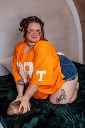 Person in bright orange university t-shirt poses on dark green velvet couch showing tattoos.
