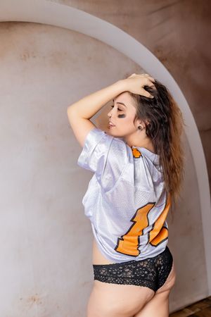 A figure in a white cropped t-shirt with orange graphics and black lace shorts poses against a textured beige wall.