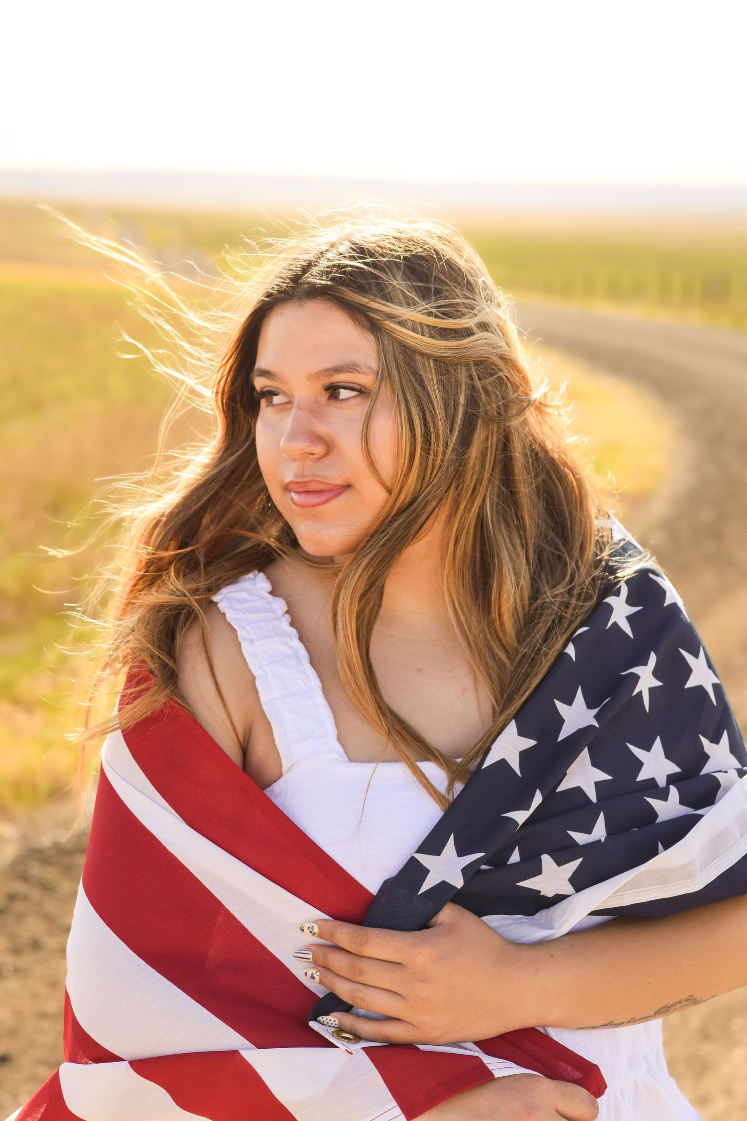 Young woman with long hair wrapped in an American flag stands on a rural dirt road at golden hour.
