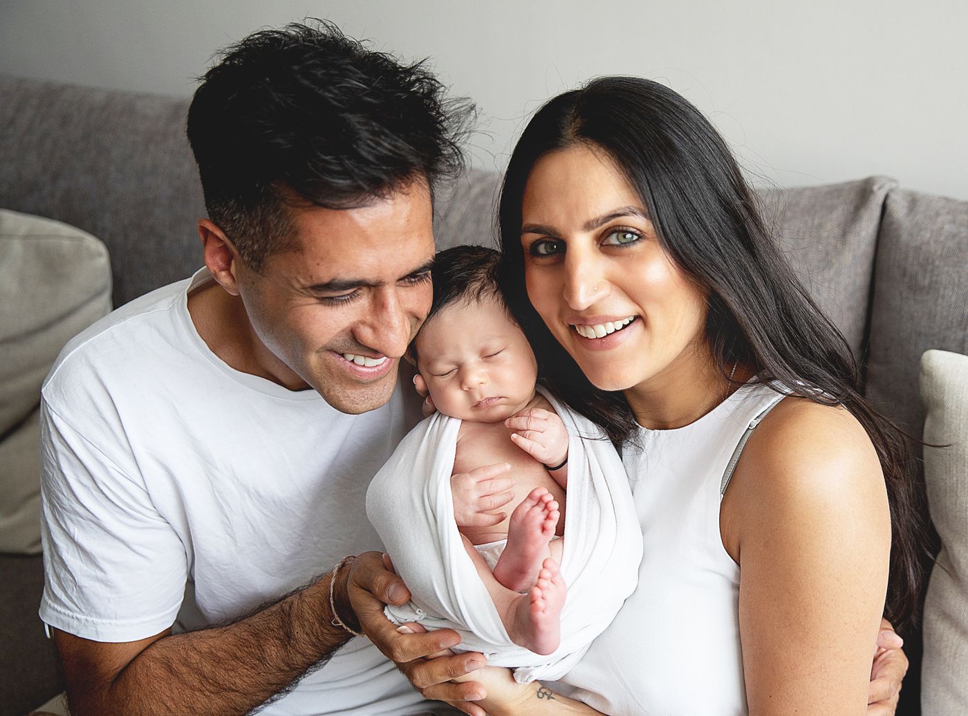 Happy couple in white shirts smiling while holding their sleeping newborn baby wrapped in white swaddle. During a homenewborn session Sidcup