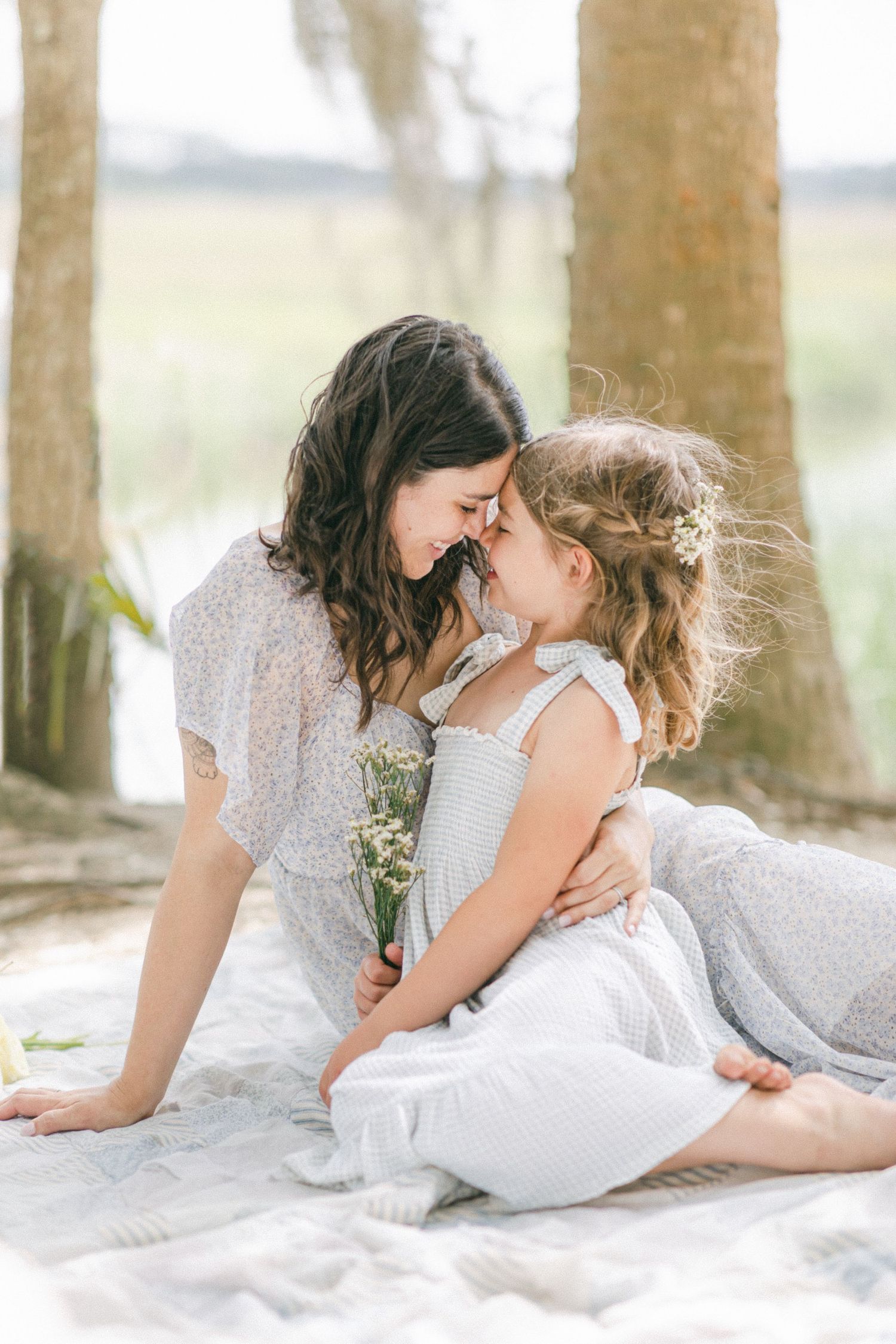 A mother and young daughter share a tender nose-to-nose moment outdoors, sitting on a blanket near trees.
