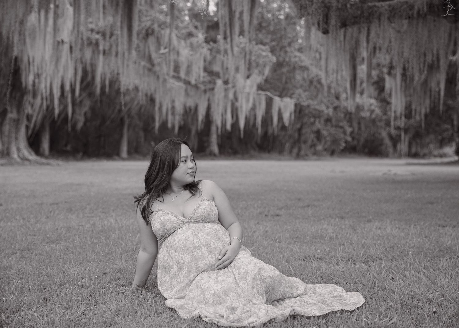 Black and white maternity photo of a pregnant woman in a floral gown sitting on grass under Spanish moss trees.