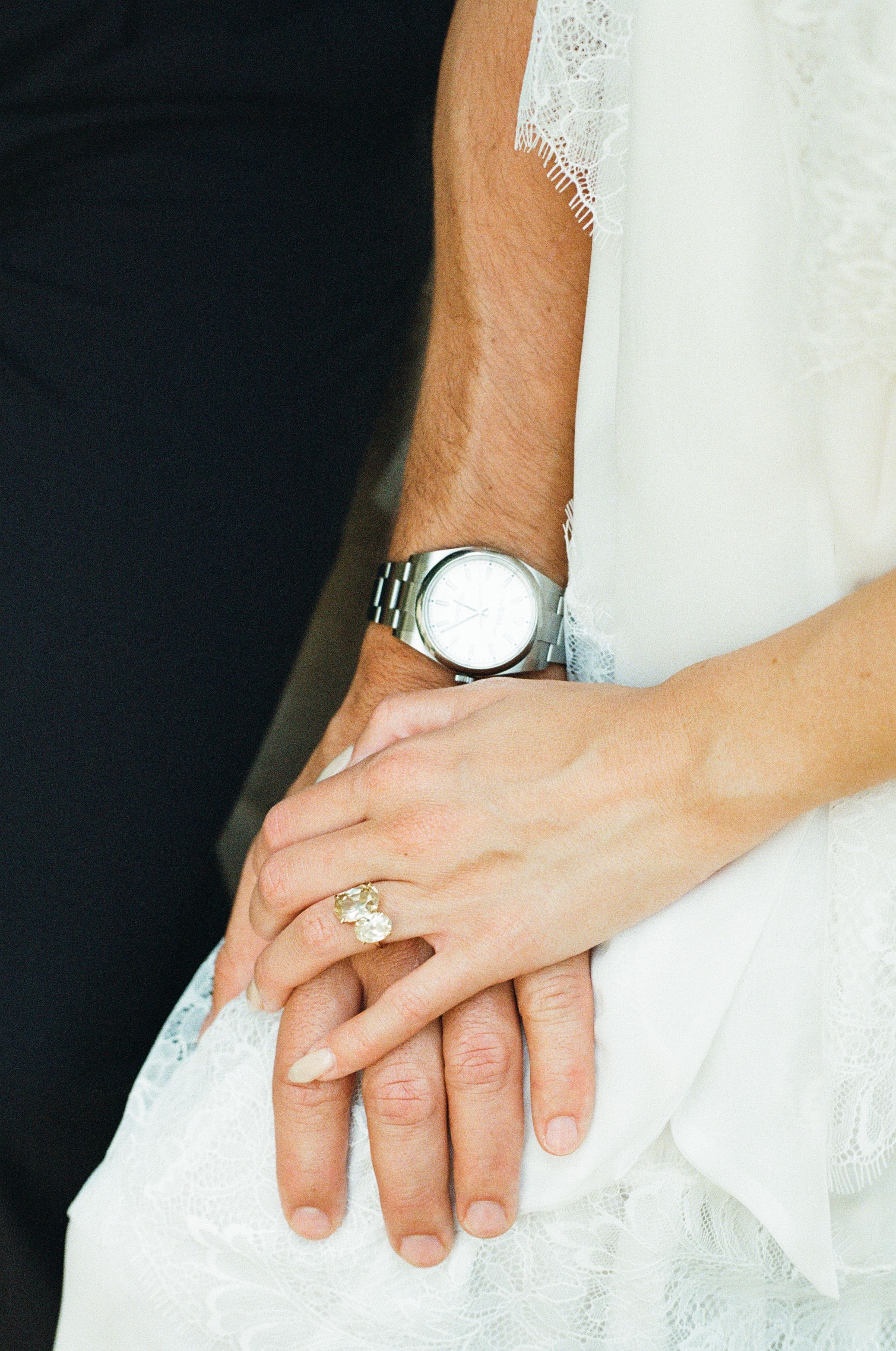 Close-up of bride and groom hands, featuring a diamond engagement ring and silver watch, resting on white lace wedding dress.