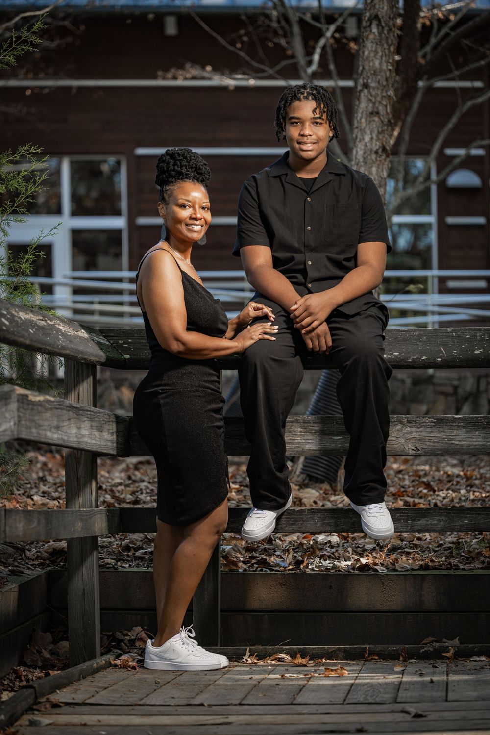 Mother and teenage son in matching black outfits posing together on wooden steps outside a building.
