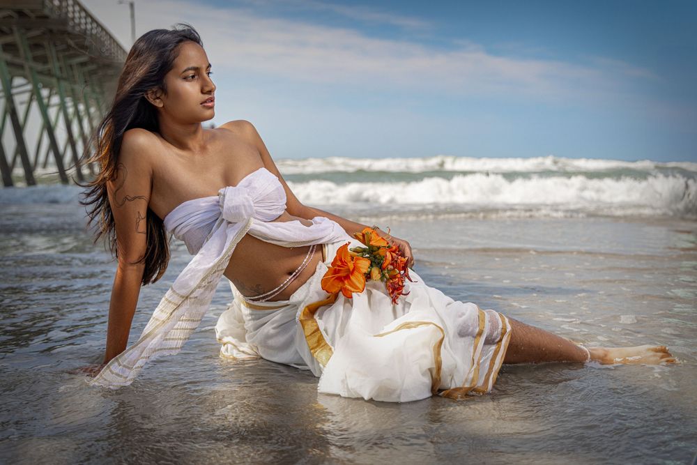Woman in white off-shoulder outfit sitting on a beach shoreline holding a bouquet of orange flowers.