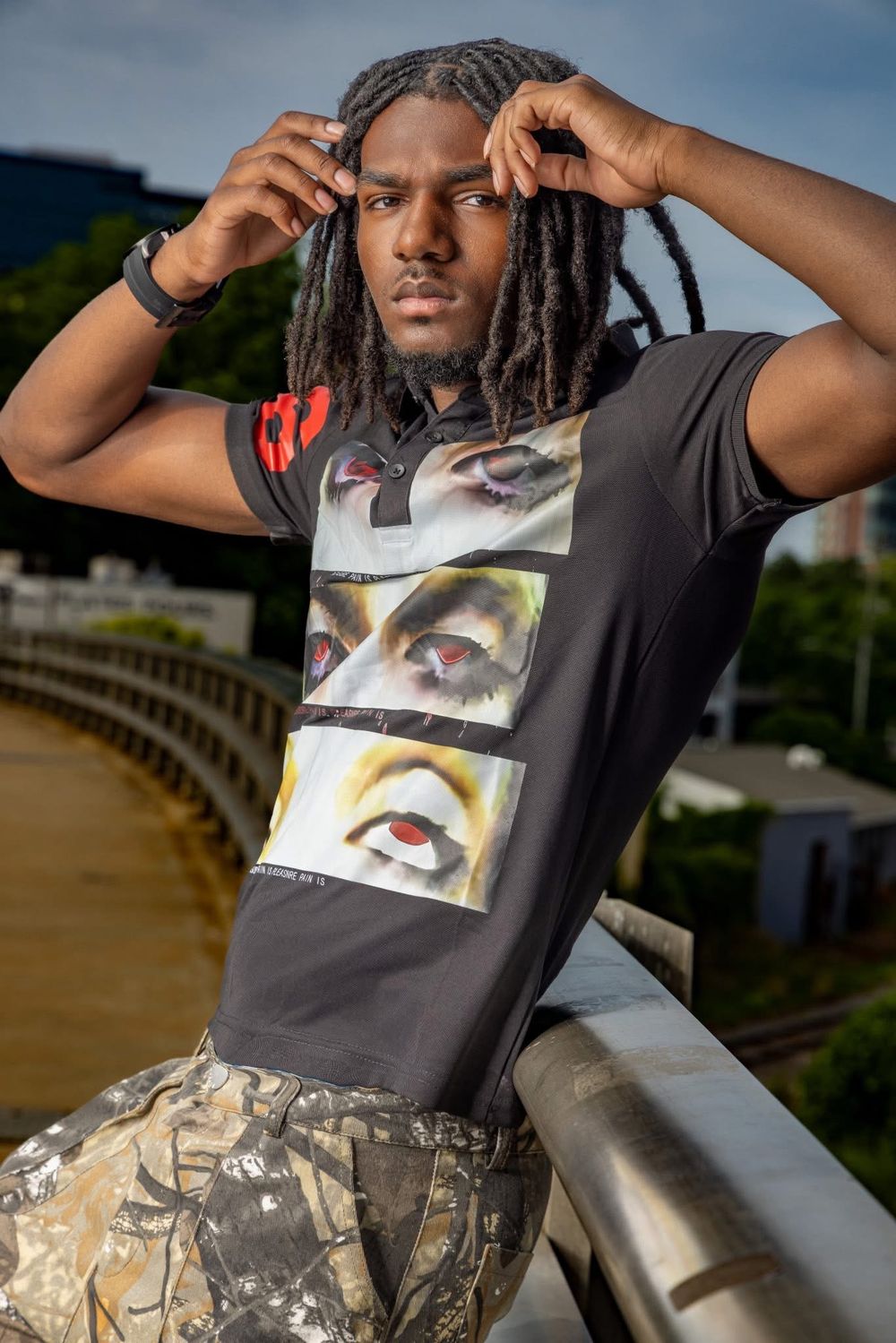 Young man with dreadlocks in graphic tee leaning on urban bridge railing.