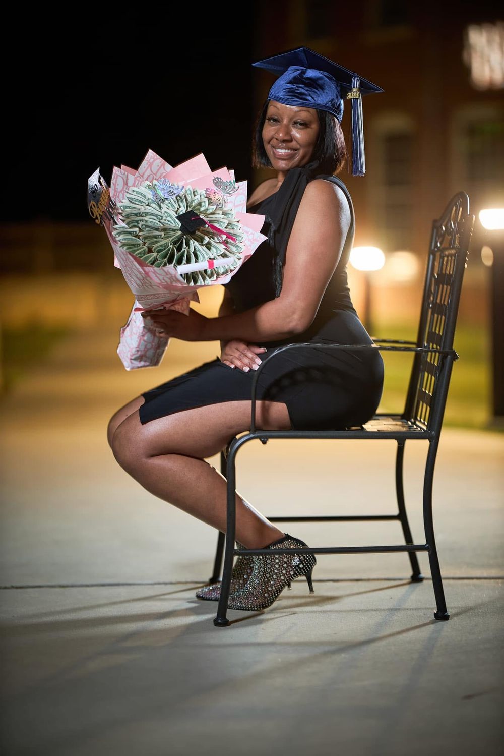Woman in graduation cap holds bouquet while seated on bench at night.