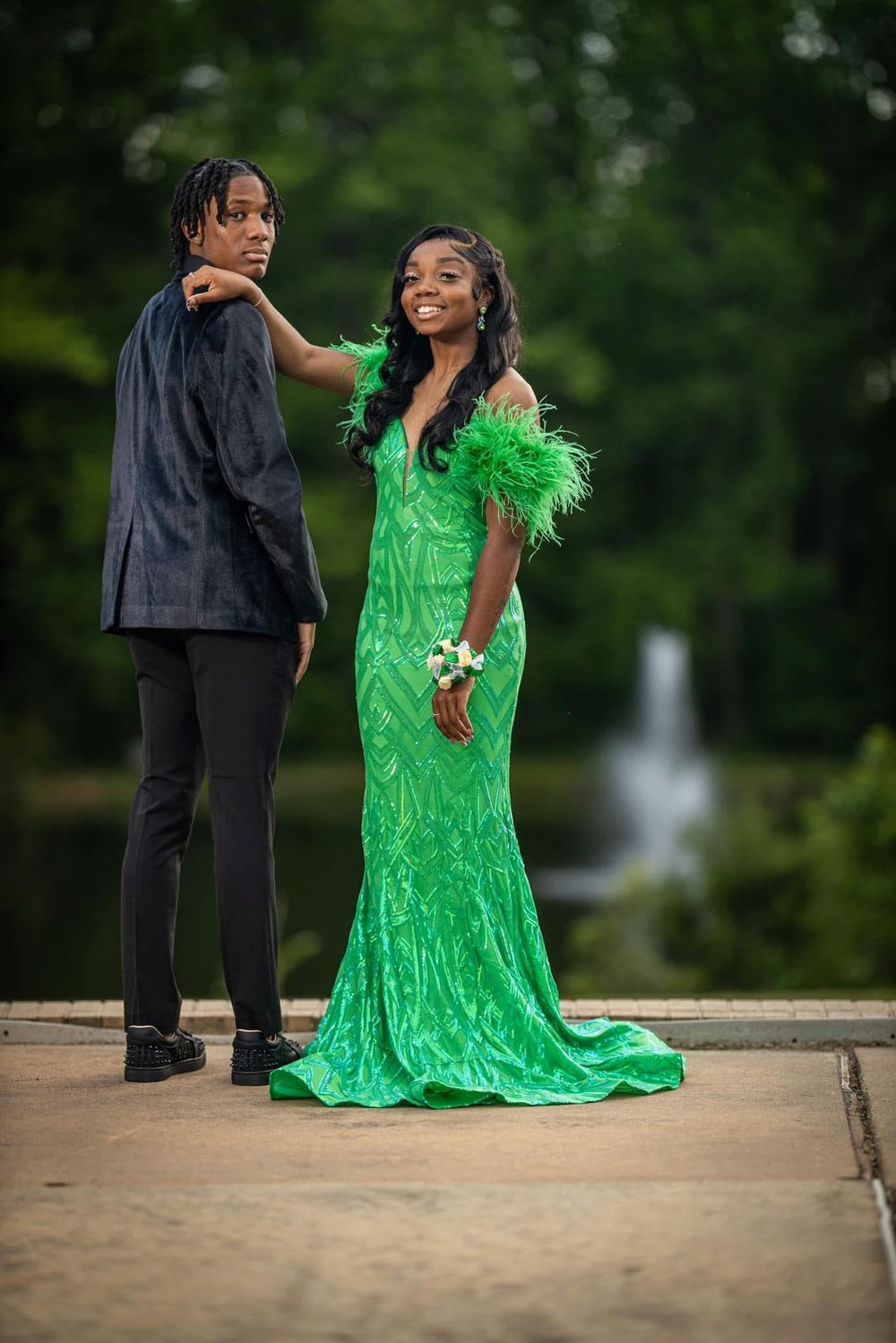 Couple posing in elegant formalwear, woman in green feathered gown near fountain.