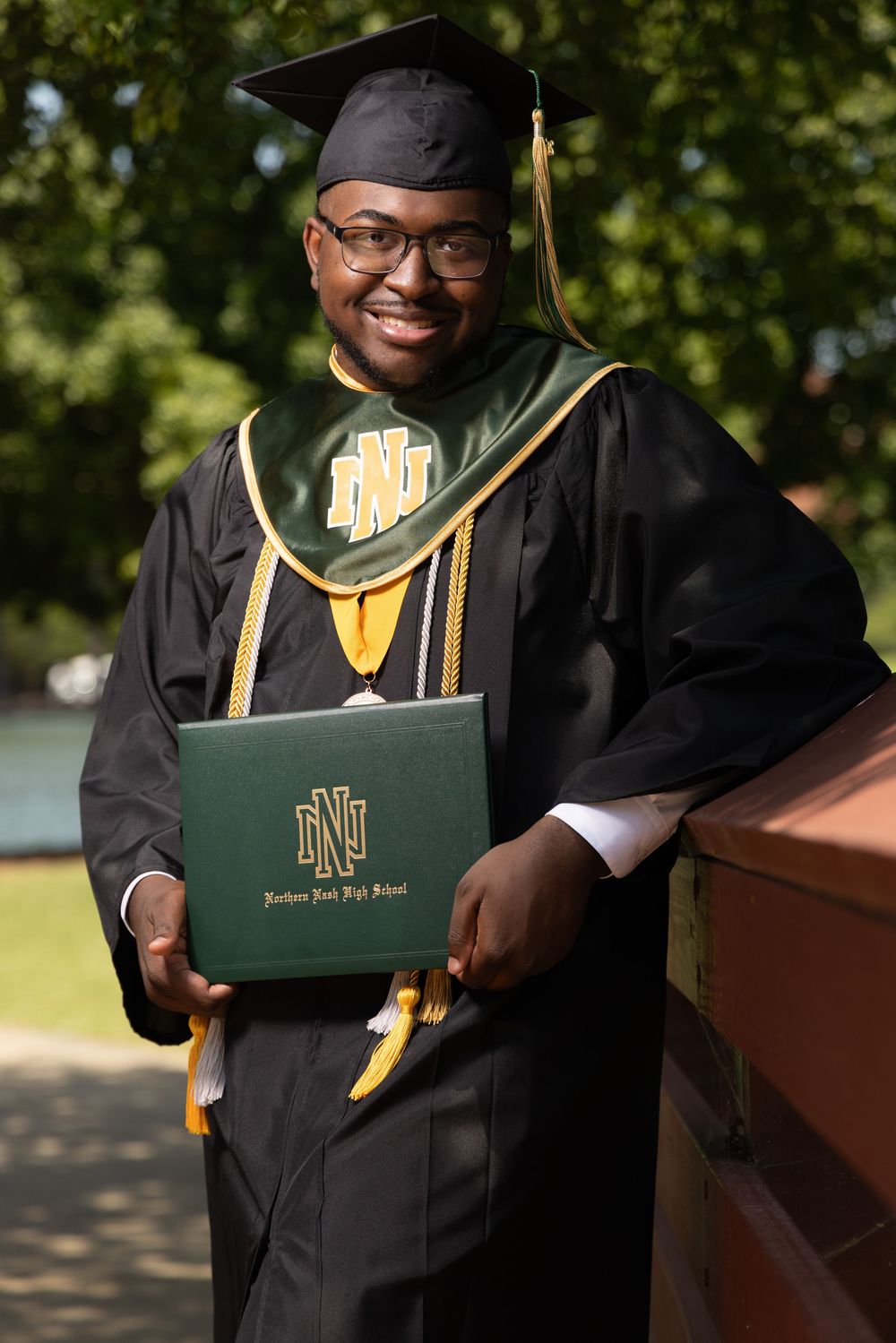 Smiling graduate in black cap and gown holding a green diploma from Northern Nash High School outdoors.