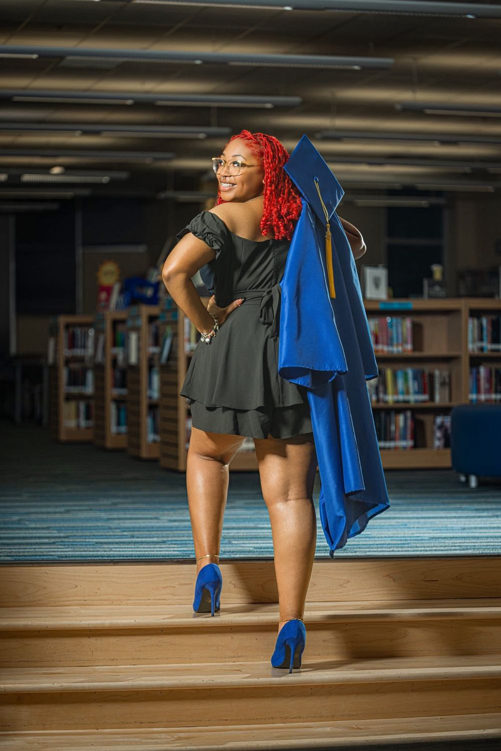 Graduate in black dress carries blue cap and gown in library.