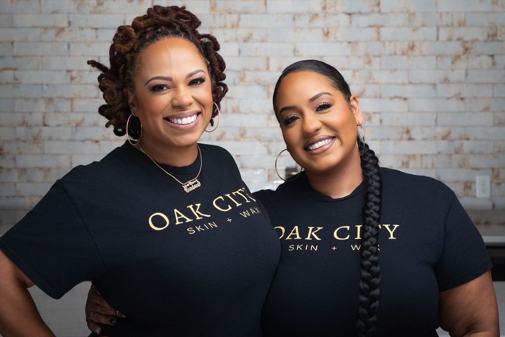 Two women smiling in matching black Oak City Skin and Wellness branded t-shirts against a white brick wall.