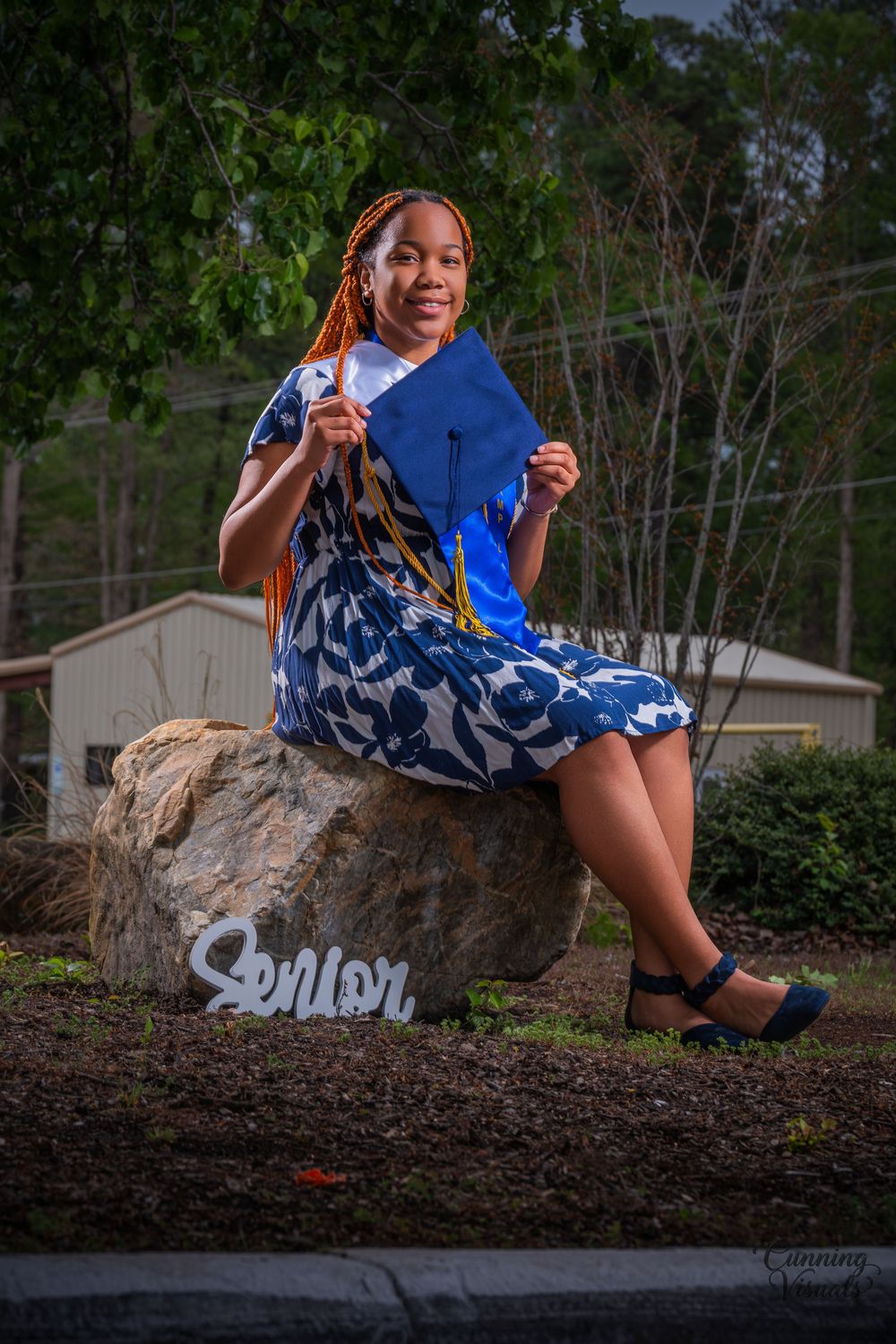 Young woman in blue floral dress holding graduation cap sitting on a rock near a Senior sign outdoors.