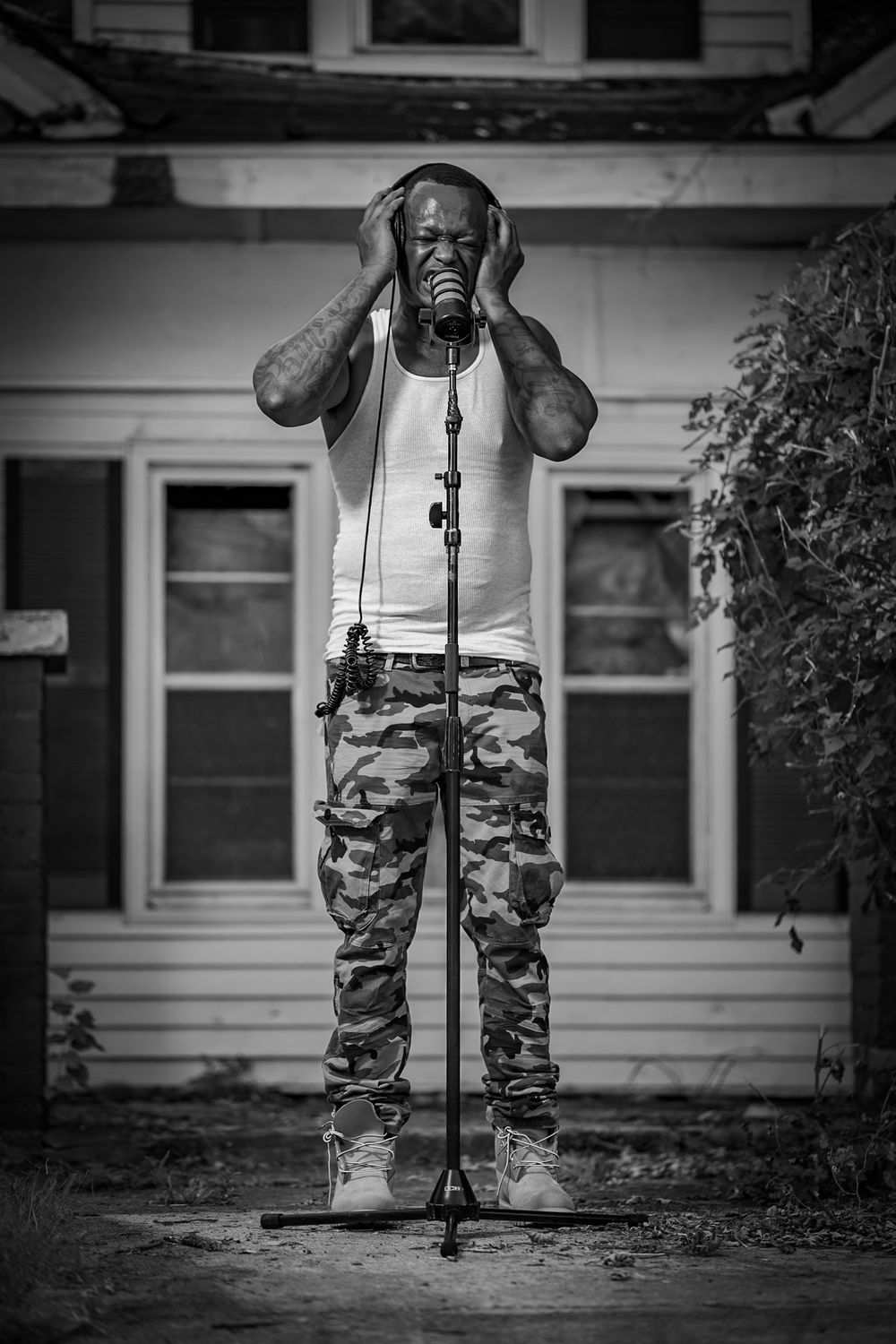Black and white portrait of a man recording at a microphone with headphones outdoors in front of a house.