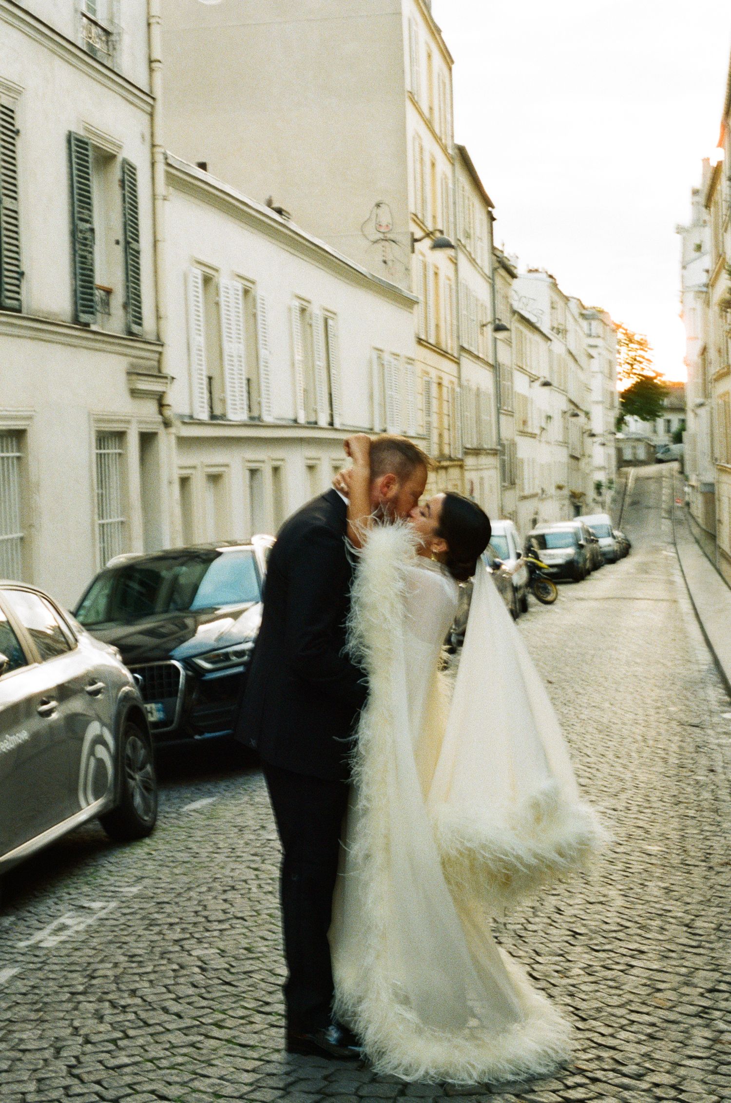 A bride and groom share a romantic kiss on a cobblestone street in Paris, France, with classic Haussmann buildings in the background.