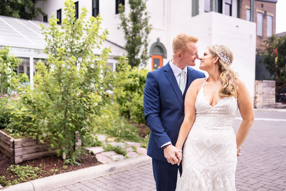 Bride and groom holding hands and gazing at each other outdoors, wearing white lace gown and navy suit.