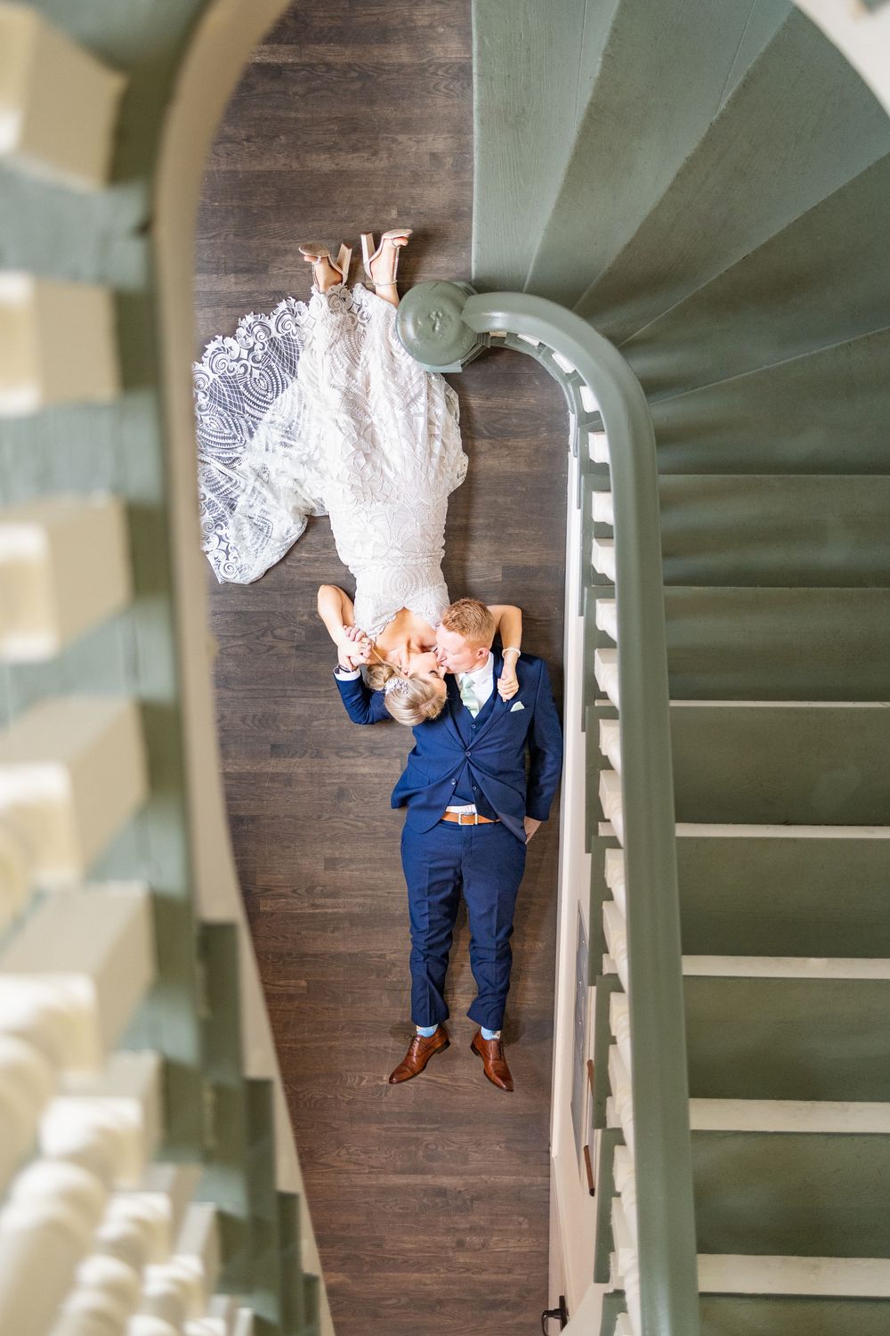 Aerial view of bride and groom sharing a kiss on a spiral staircase landing, bride in lace gown, groom in navy suit.