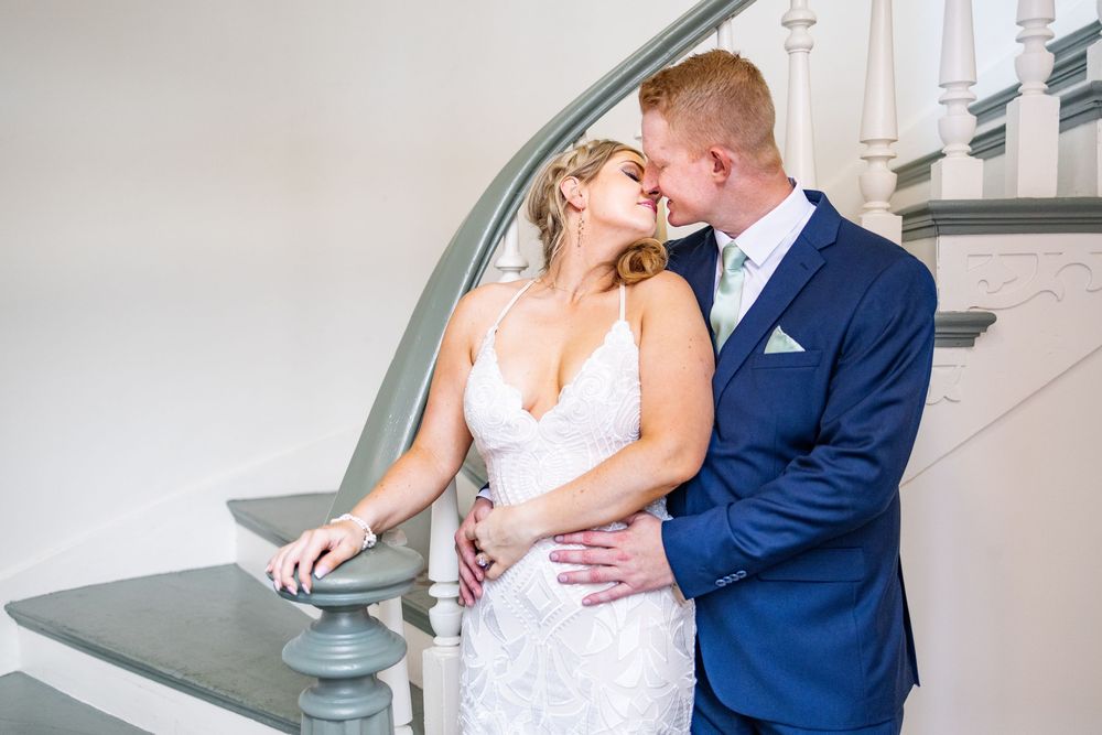 Bride and groom share a romantic kiss near a white staircase, bride in lace gown, groom in navy suit.