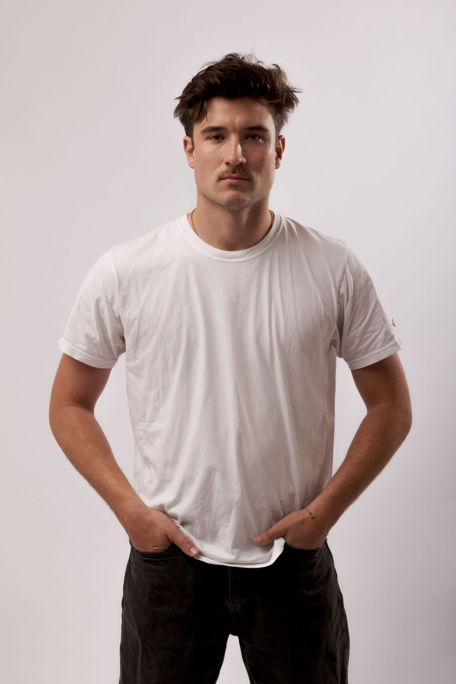 Young man with mustache wearing a white t-shirt and dark jeans, posing with hands in pockets against a light background.