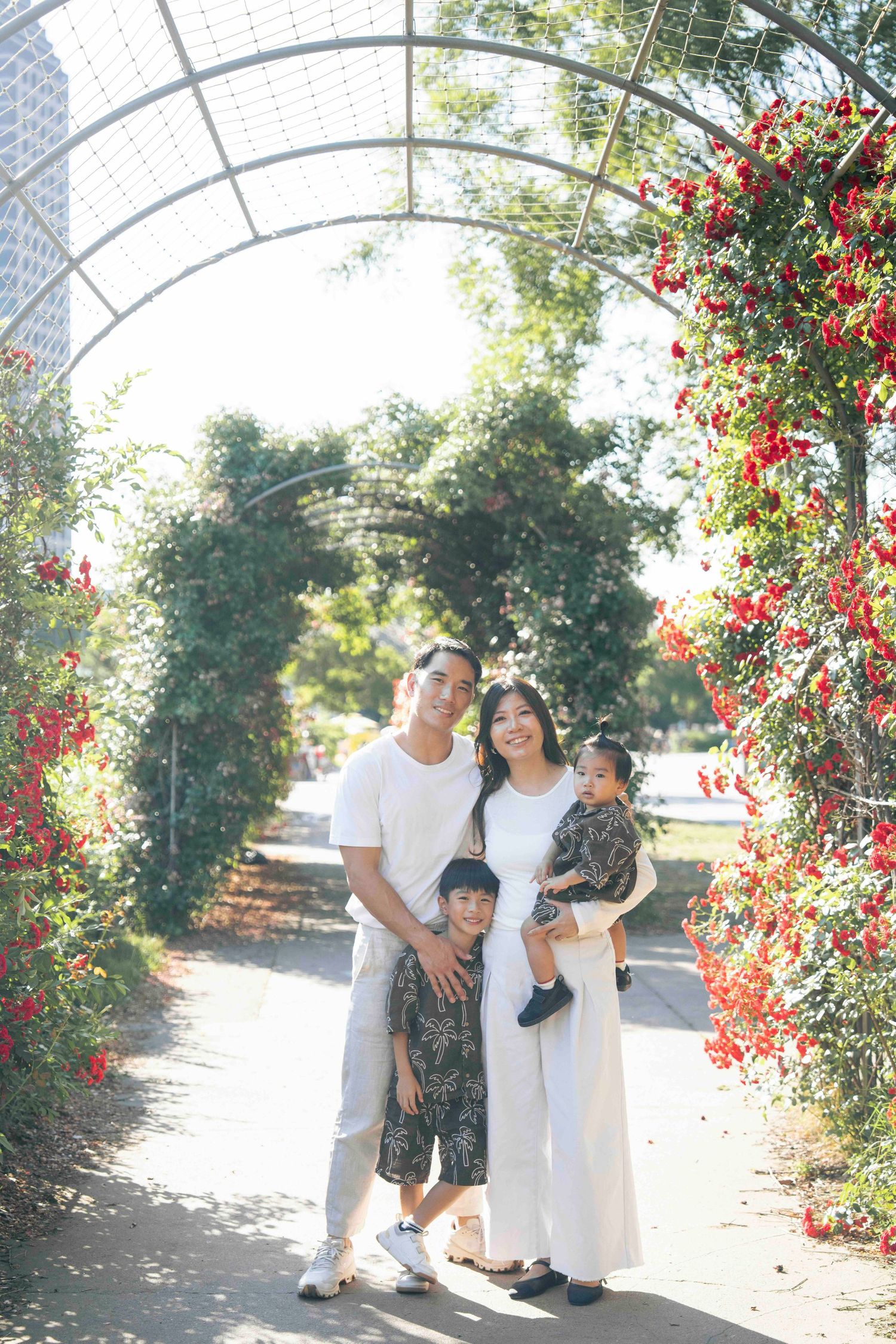 Family of four posing under a floral arch with red blooms in a sunlit garden park.