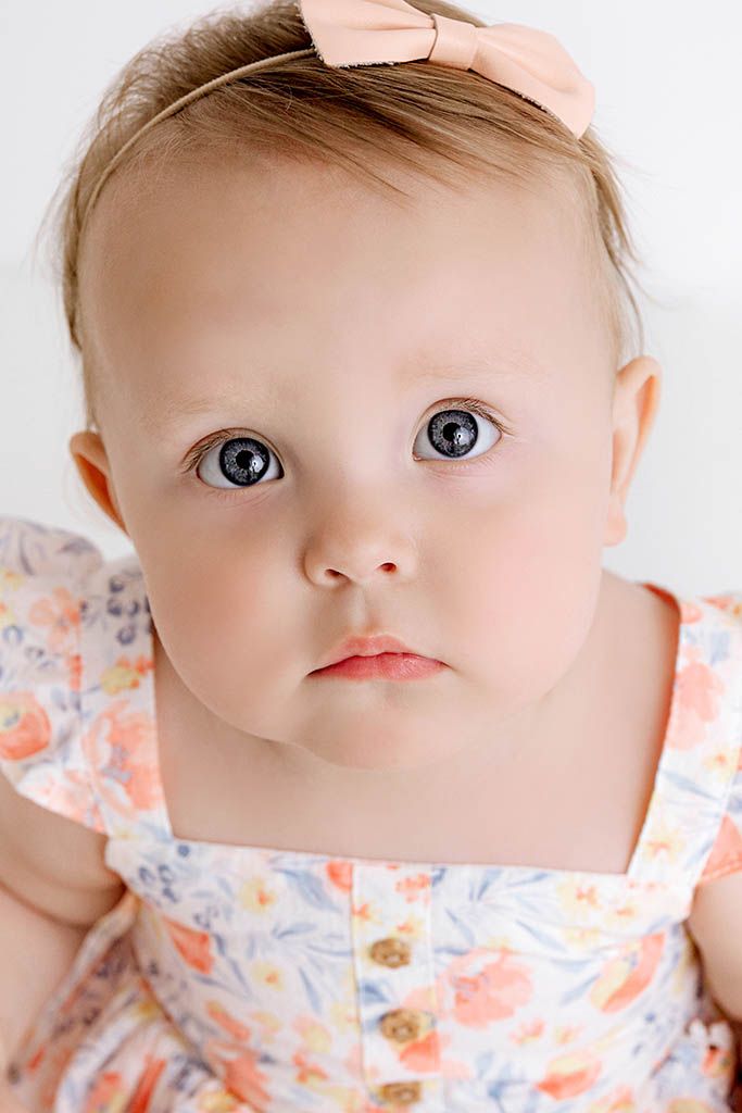 Adorable baby girl with blue eyes wearing a floral dress and peach bow headband.