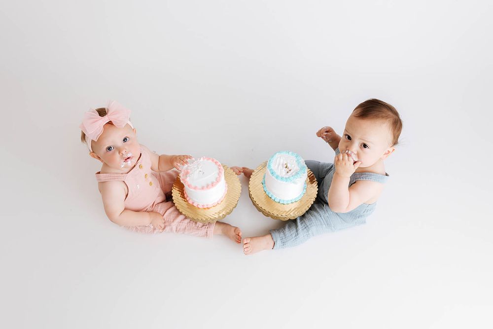 Two babies in pink and blue outfits sit facing each other with matching first birthday smash cakes on white background.