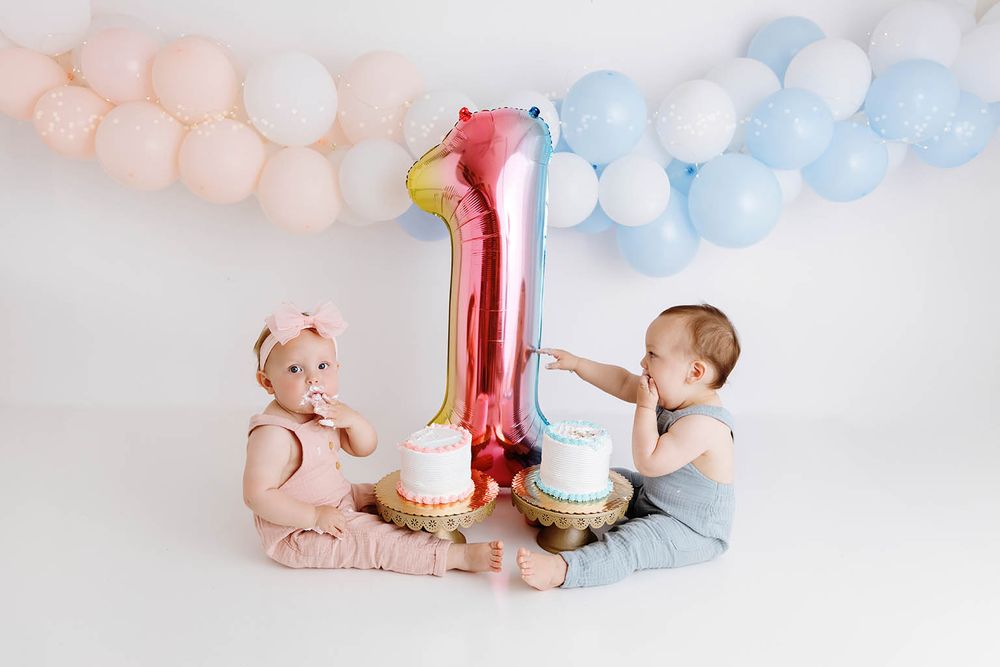 Twin babies celebrate their first birthday with smash cakes and a number one balloon at a white studio photo shoot.
