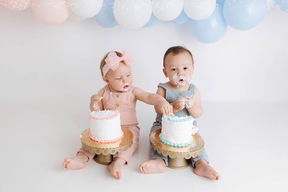 Twin babies in pink and blue outfits smash their first birthday cakes on gold stands with balloon backdrop.