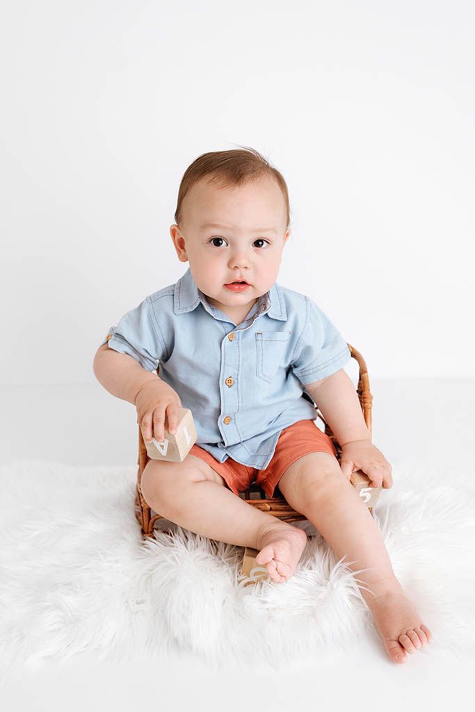 A cute baby boy in a blue shirt and orange shorts sits in a wicker chair holding wooden blocks on a white fur rug.