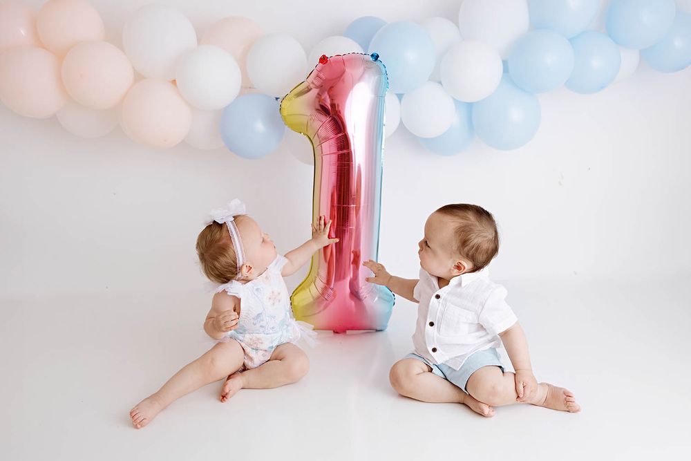 Twin babies celebrating first birthday with a large number one balloon and pastel decor.