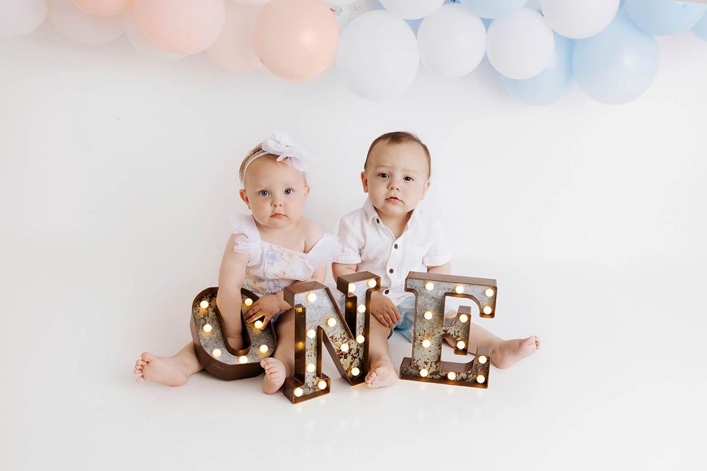 Twin babies sit together at their first birthday photo shoot with lit ONE marquee letters and pink and blue balloons.
