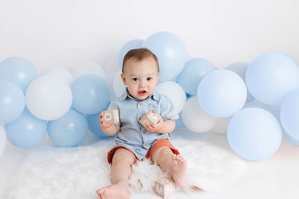 Baby boy sitting on white fur rug holding gifts surrounded by blue balloons.