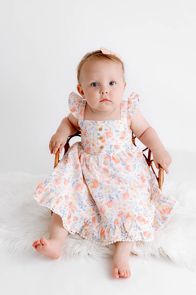 Adorable baby girl in floral ruffle dress with pink bow headband sits on fur rug for portrait photo.