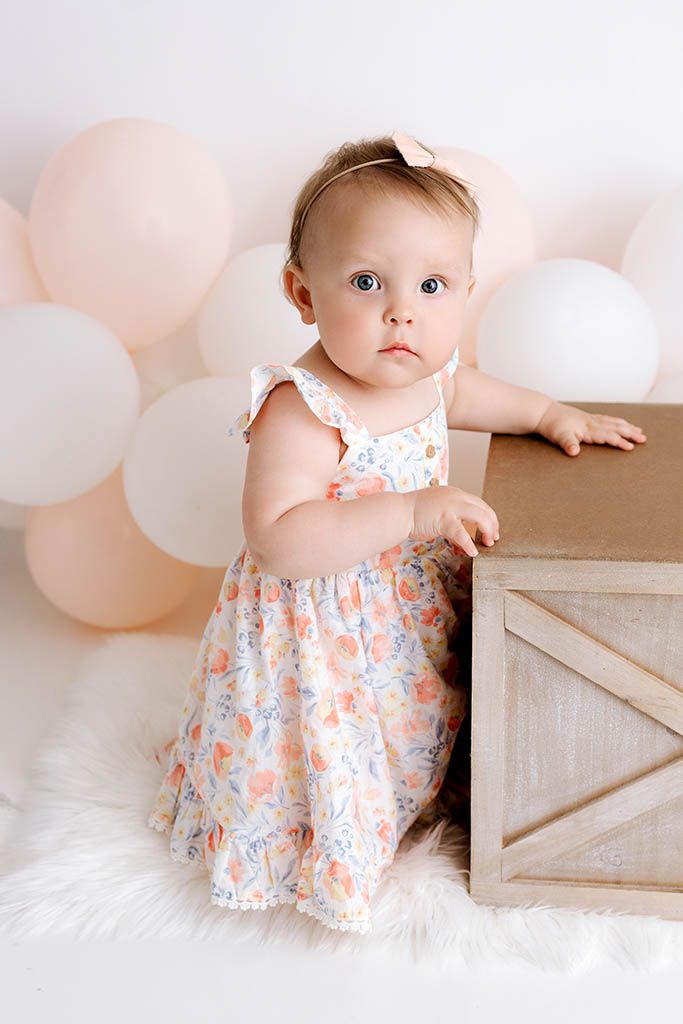 Baby girl in floral dress standing by wooden crate surrounded by peach balloons.