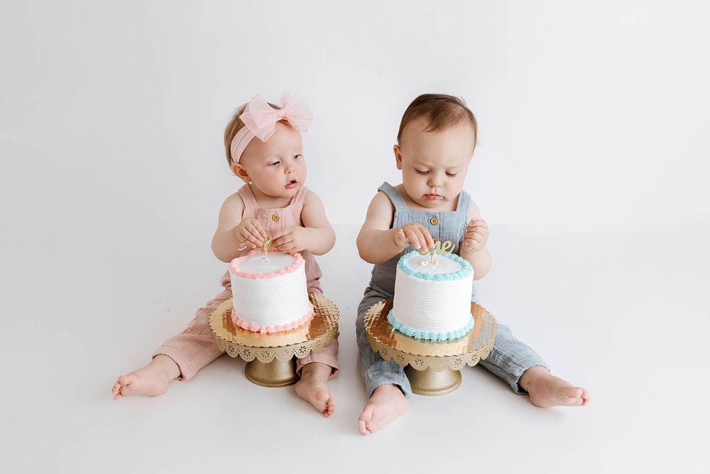 Twin boy and girl babies in coordinating outfits explore their white first birthday smash cakes on gold stands.