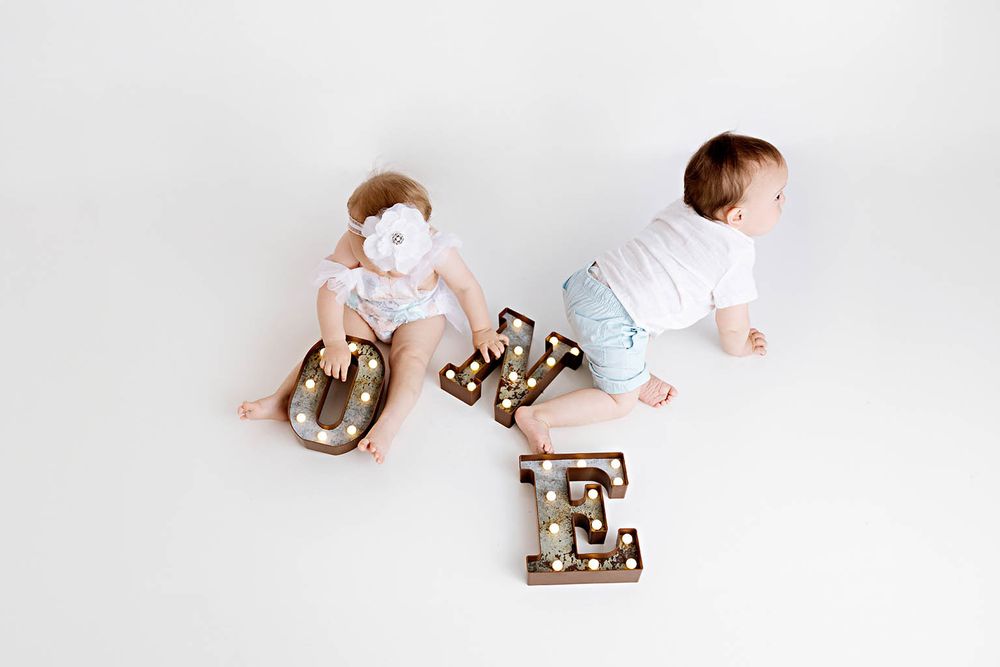 Two babies sit back-to-back on white floor with decorative marquee letters spelling LOVE between them.