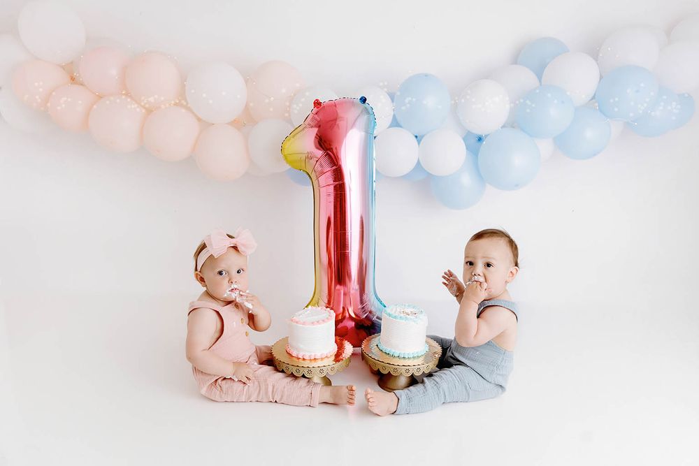 Twin babies celebrate first birthday with smash cakes and a number one balloon surrounded by pink and blue balloons.