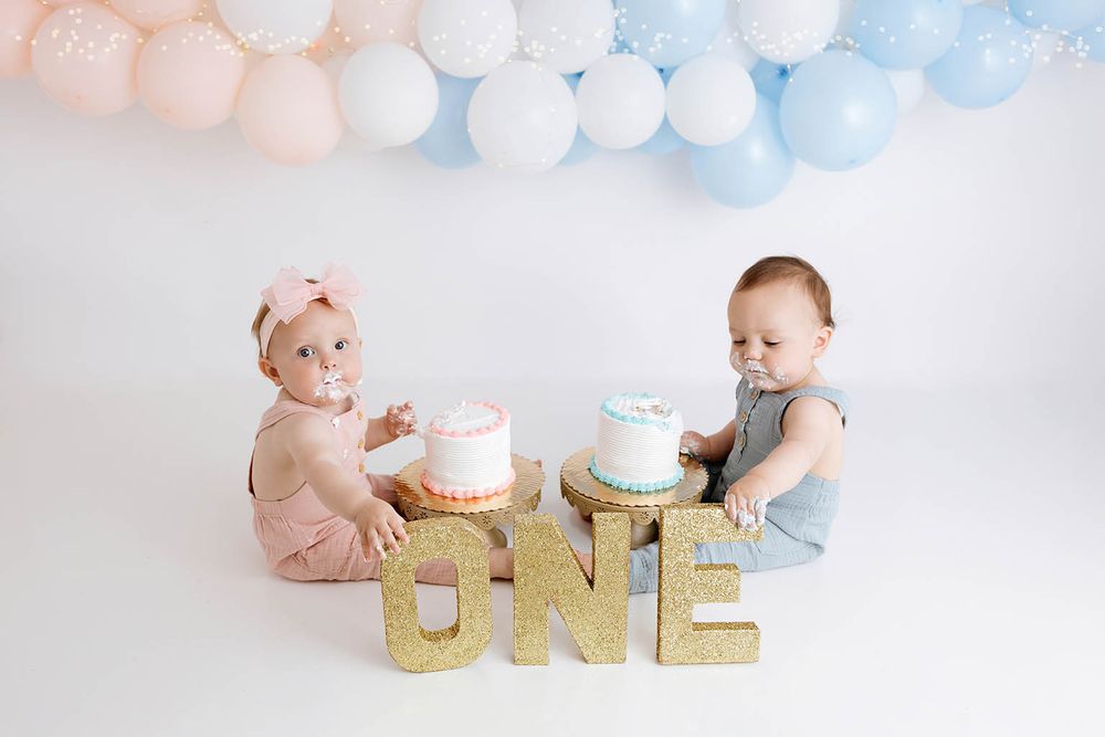 Twin babies celebrate first birthday smashing cakes beside gold glitter ONE sign with pink and blue balloon garland.
