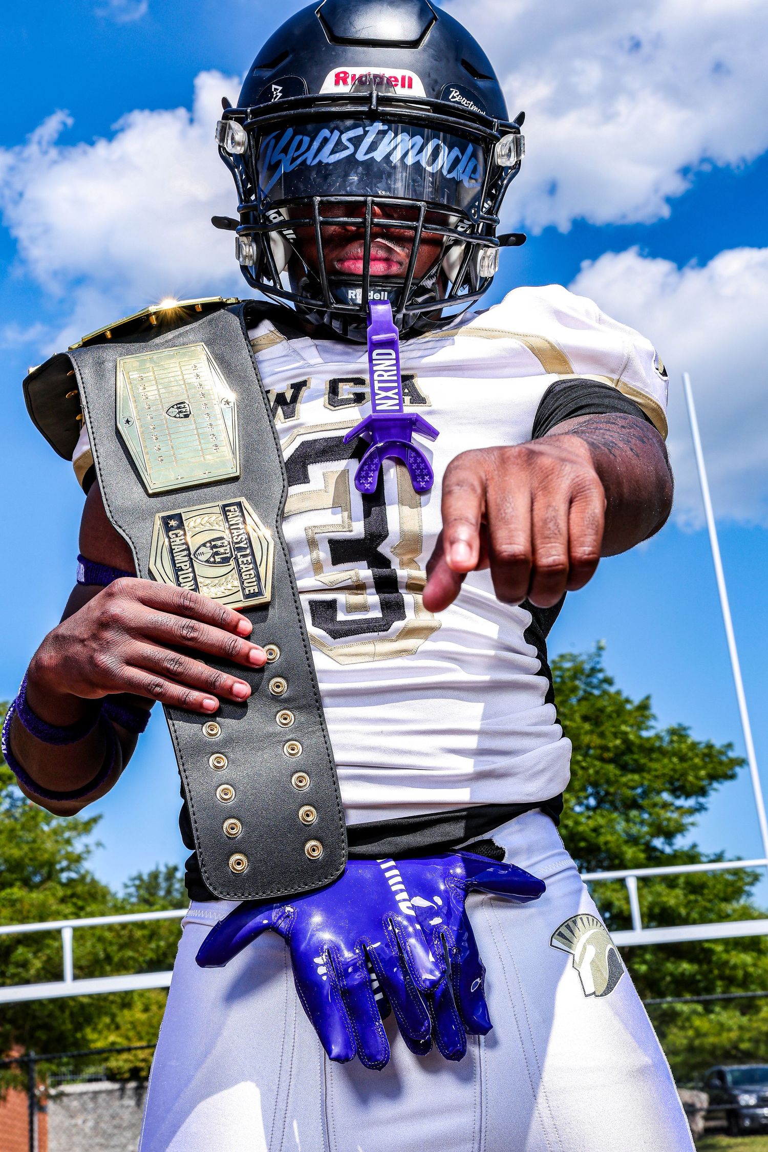 Football player in Beastmode helmet holds championship belt, pointing at camera in white and purple uniform outdoors.