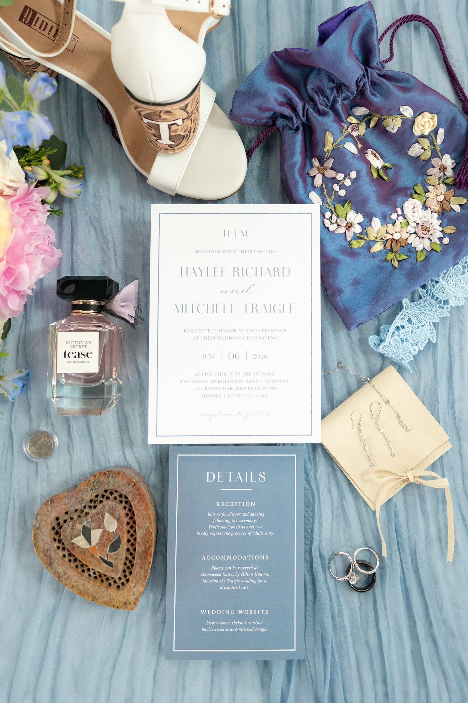 Elegant wedding invitation suite with details card, perfume, rings, floral bag, and bridal shoes on a soft blue background.