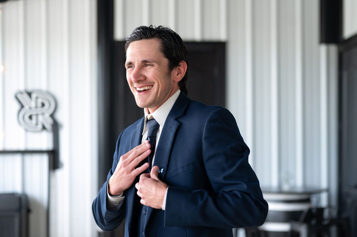 Smiling man in navy blue suit adjusting tie, standing in modern venue with industrial decor.
