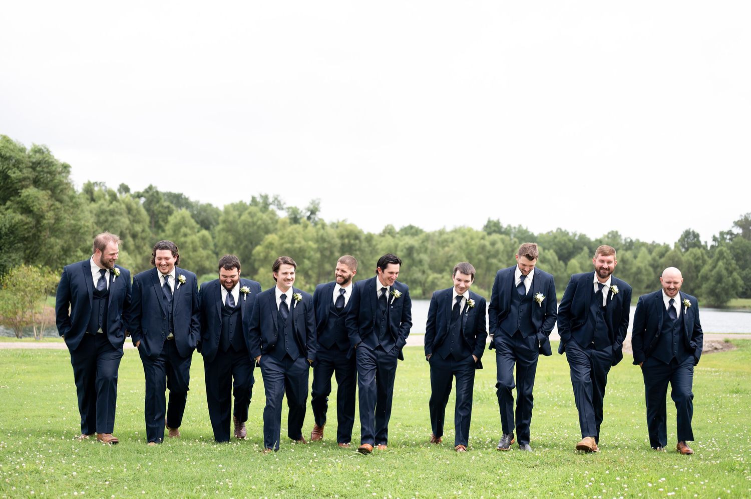 Groomsmen in navy blue suits walking together on green grass at an outdoor wedding ceremony.