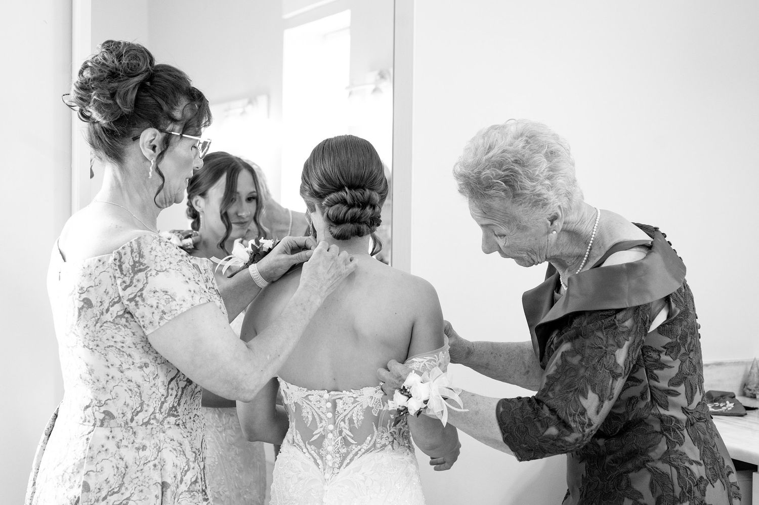 Black and white photo of bride being helped into wedding dress by family members, reflecting in mirror.